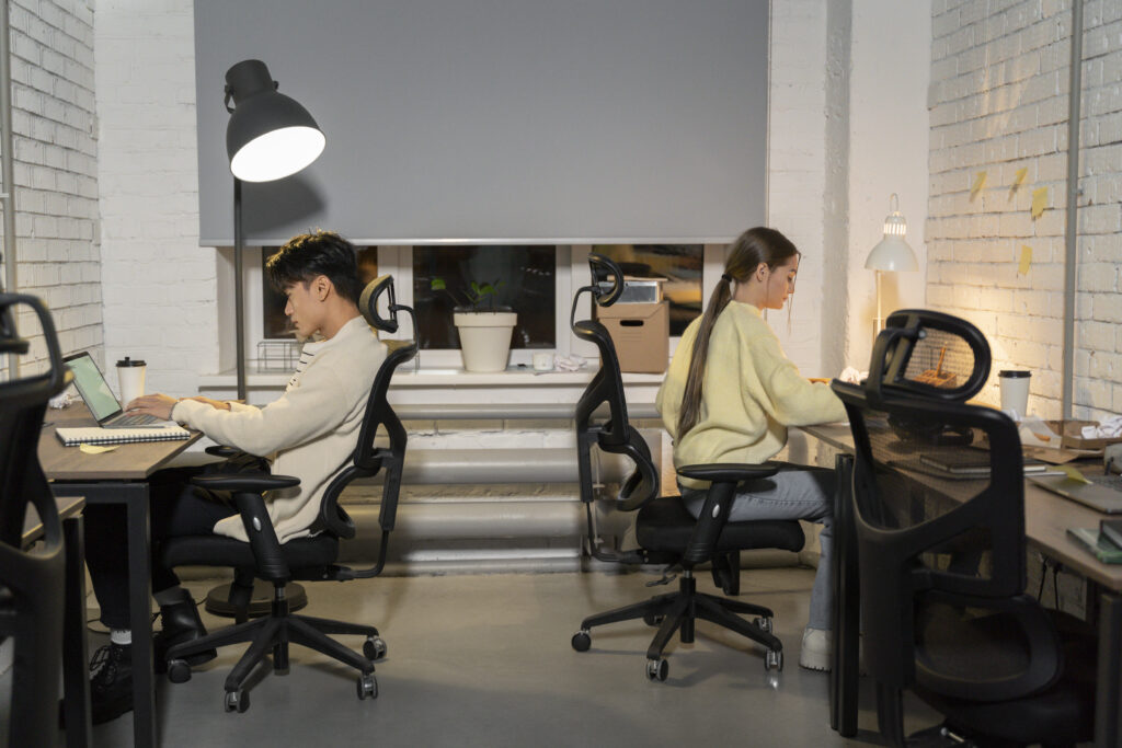 Two professionals working back-to-back at desks in a modern office, using ergonomic office chairs and laptops under desk lamps.