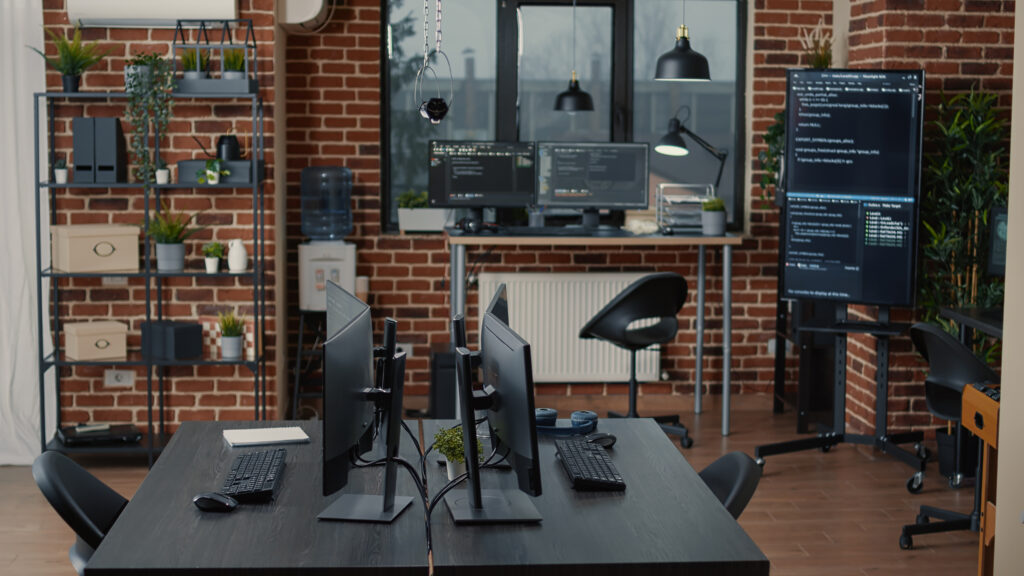 Modern IT office workspace with multiple computer desks, dual monitors displaying programming code, ergonomic chairs, and brick wall interior.